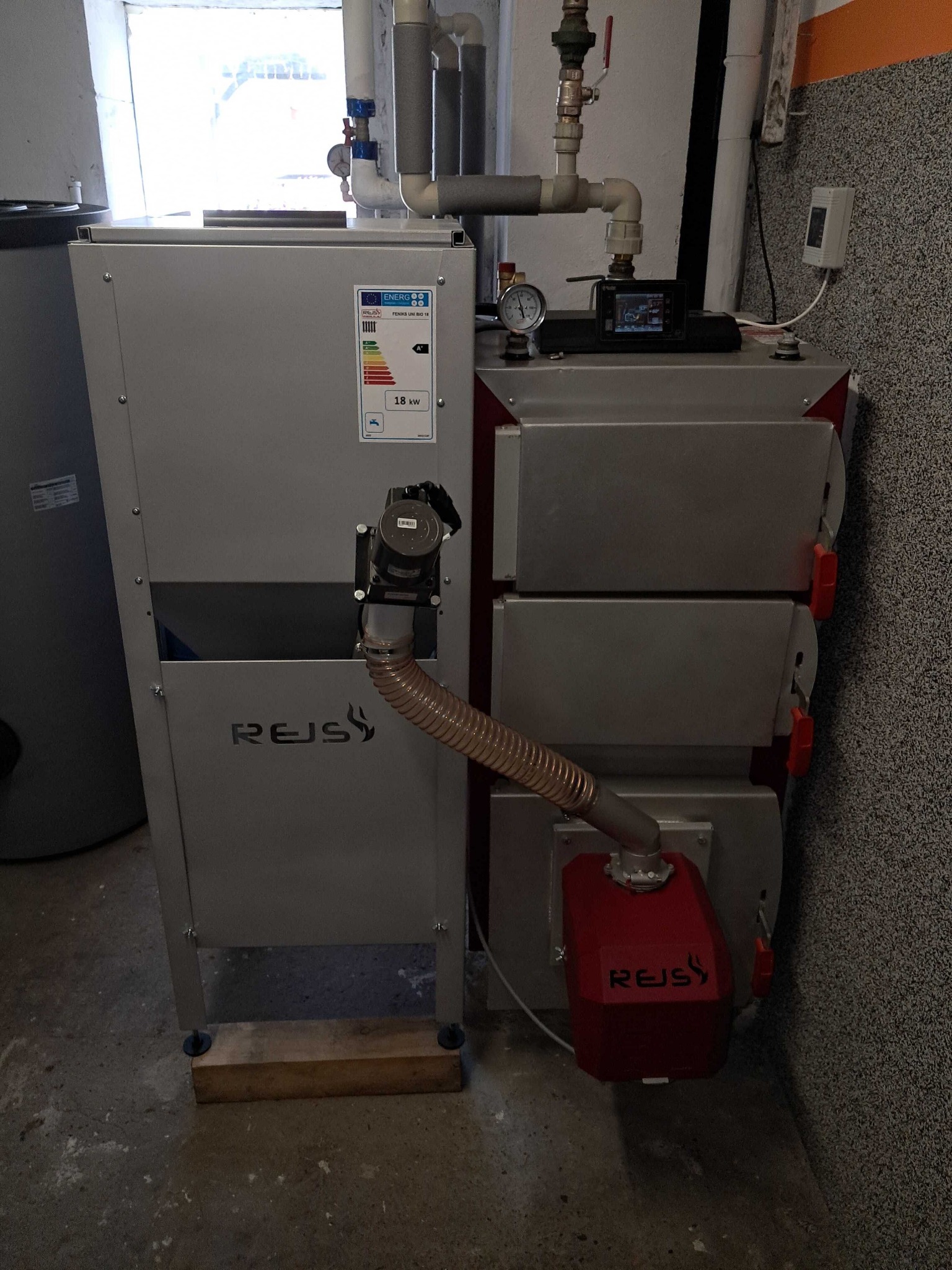 Szary piec marki REJS z podajnikiem, widoczna etykieta energetyczna, manometr i sterownik elektroniczny na górze, rura doprowadzająca paliwo z podajnika do pieca, ustawiony na drewnianych klockach...