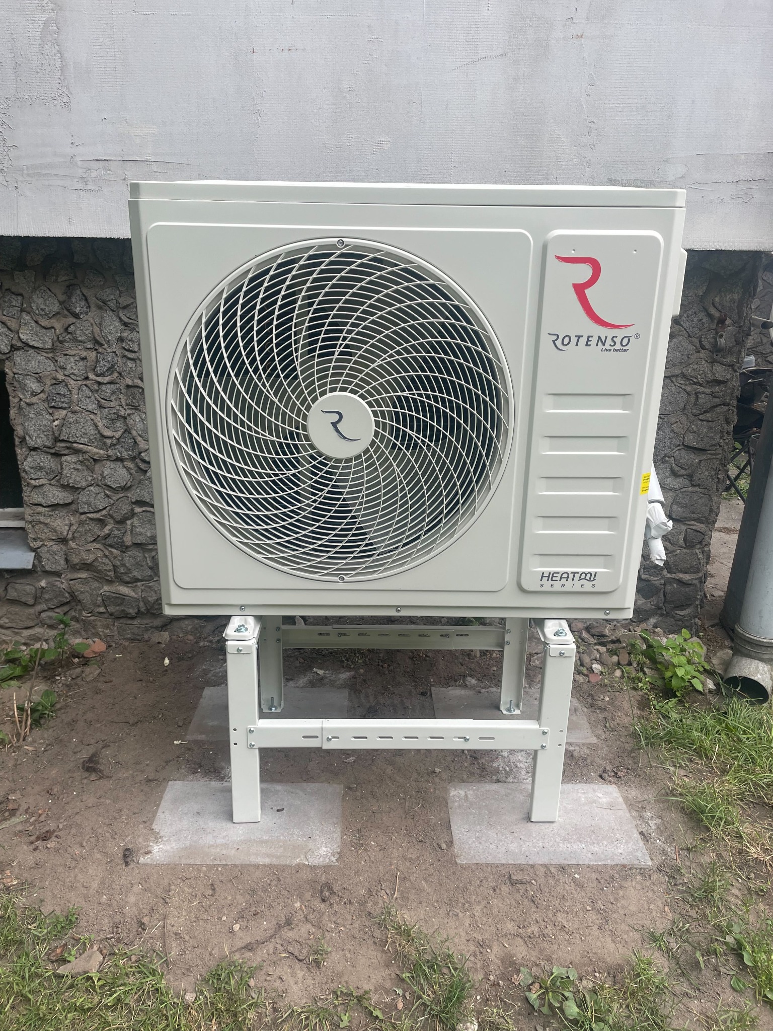 Zewnętrzna jednostka pompy ciepła marki Rotenso, zamontowana na stalowej konstrukcji na zewnątrz budynku z kamienną elewacją i oknem w tle.
