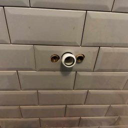 BSOLUTIONS KRZYSZTOF BULBAK - Ściana wyłożona szarymi, prostokątnymi płytkami typu metro z zamontowanymi, mosiężnymi króćcami i białym, plastikowym elementem instalacji wodnej.