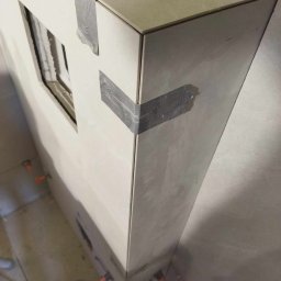 BSOLUTIONS KRZYSZTOF BULBAK - Fragment obudowy z jasnych płytek, zabezpieczonych taśmą malarską na krawędziach, w trakcie prac wykończeniowych, z widocznymi krzyżykami dystansowymi na podłodze.