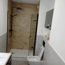 BSOLUTIONS KRZYSZTOF BULBAK - Nowoczesna łazienka po remoncie z kabiną prysznicową typu walk-in z czarnymi ramami i deszczownicą, drewnopodobnymi płytkami na ścianach w strefie prysznica oraz podwieszaną toaletą.