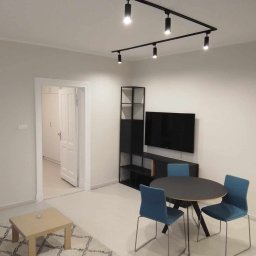 BSOLUTIONS KRZYSZTOF BULBAK - Jasne, pomalowane na szaro mieszkanie z czarnym oświetleniem punktowym, meblami i telewizorem na ścianie. W tle widoczne otwarte drzwi do innego pomieszczenia.