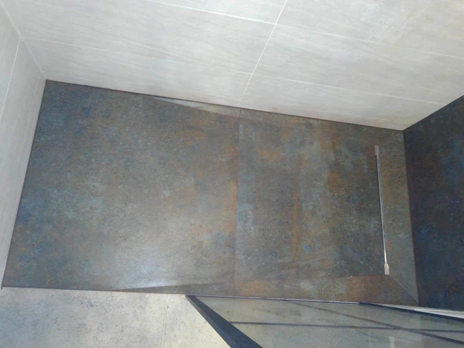 Wnętrze kabiny prysznicowej z ciemnymi, metalicznymi płytkami na podłodze i jasnymi, beżowymi płytkami na ścianach, widoczny odpływ liniowy i fragment szklanej ścianki.