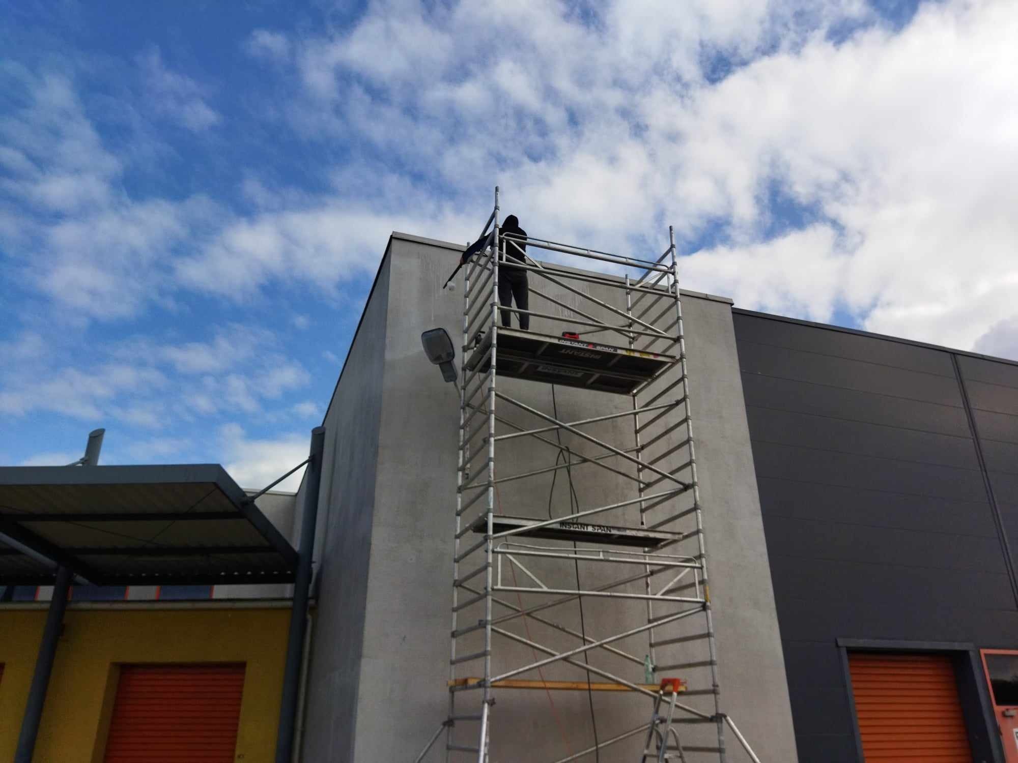 Pracownik w czarnym ubraniu maluje elewację budynku z użyciem rusztowania, widoczne niebo z chmurami.