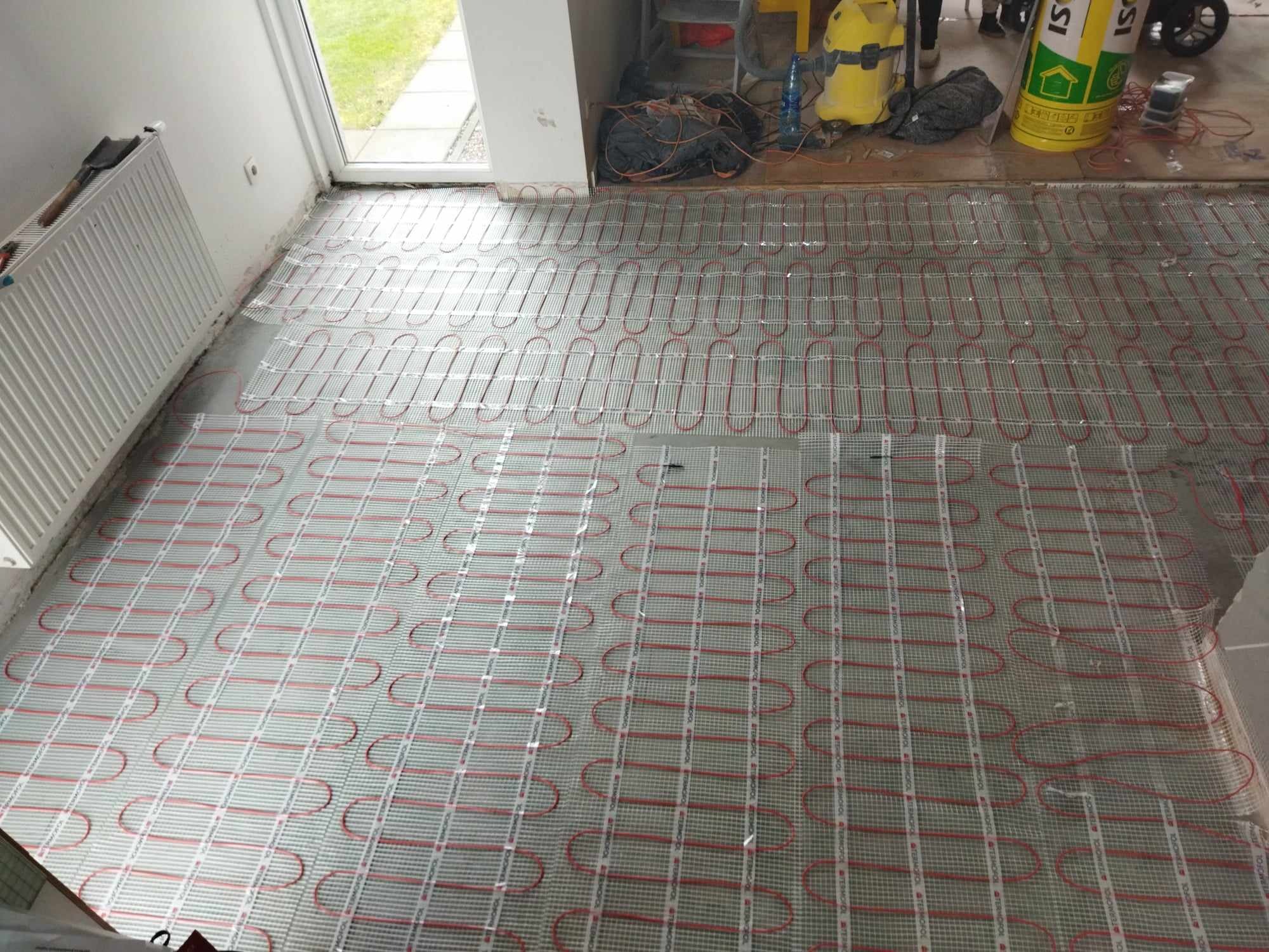 Montaż ogrzewania podłogowego: czerwone kable grzewcze ułożone na siatce, widoczne narzędzia i materiały budowlane w tle.