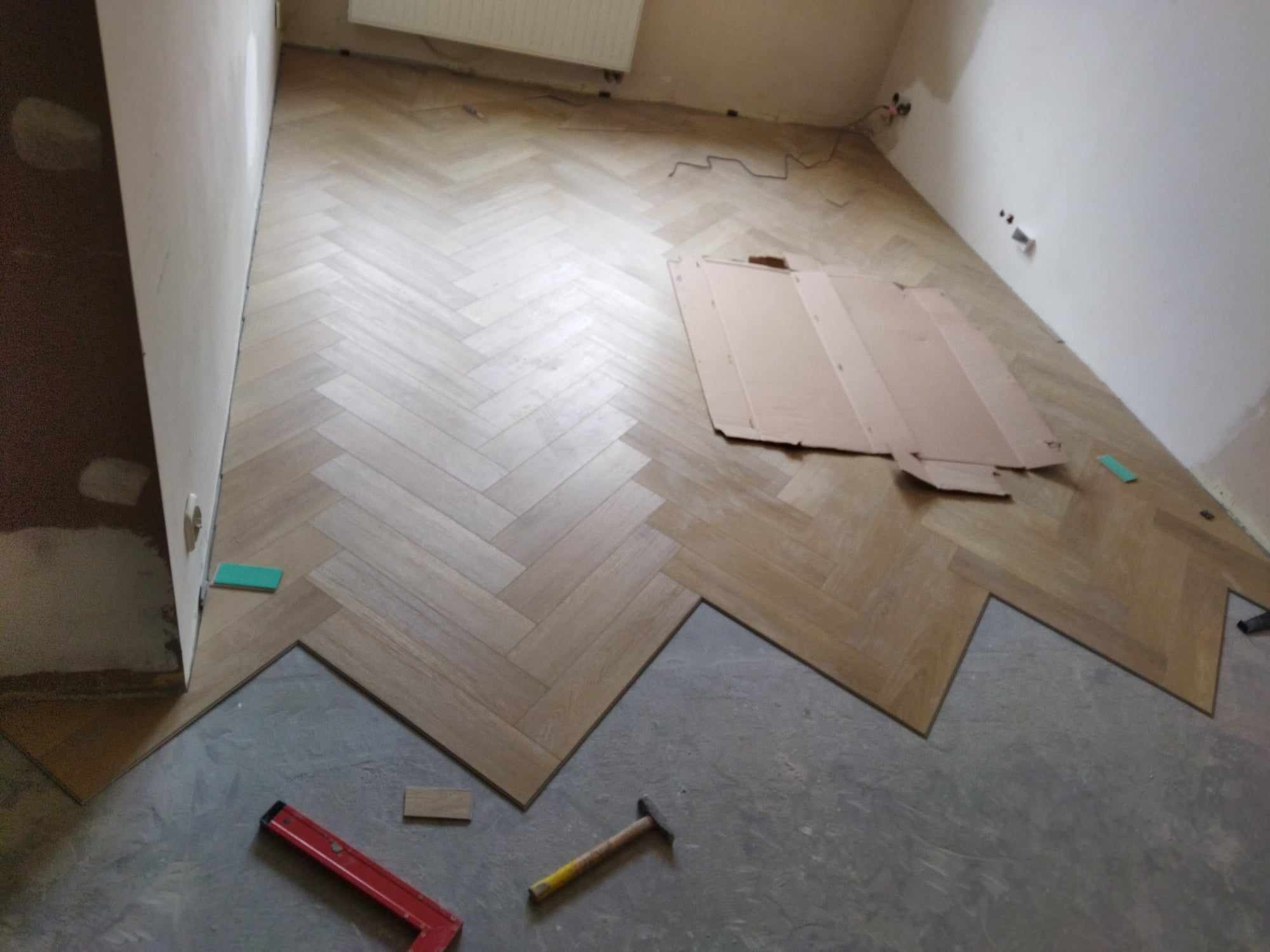 Układanie parkietu w jodełkę, widoczny fragment podłogi przed położeniem desek, poziomica, młotek i kartonowe zabezpieczenie na ułożonej części podłogi.