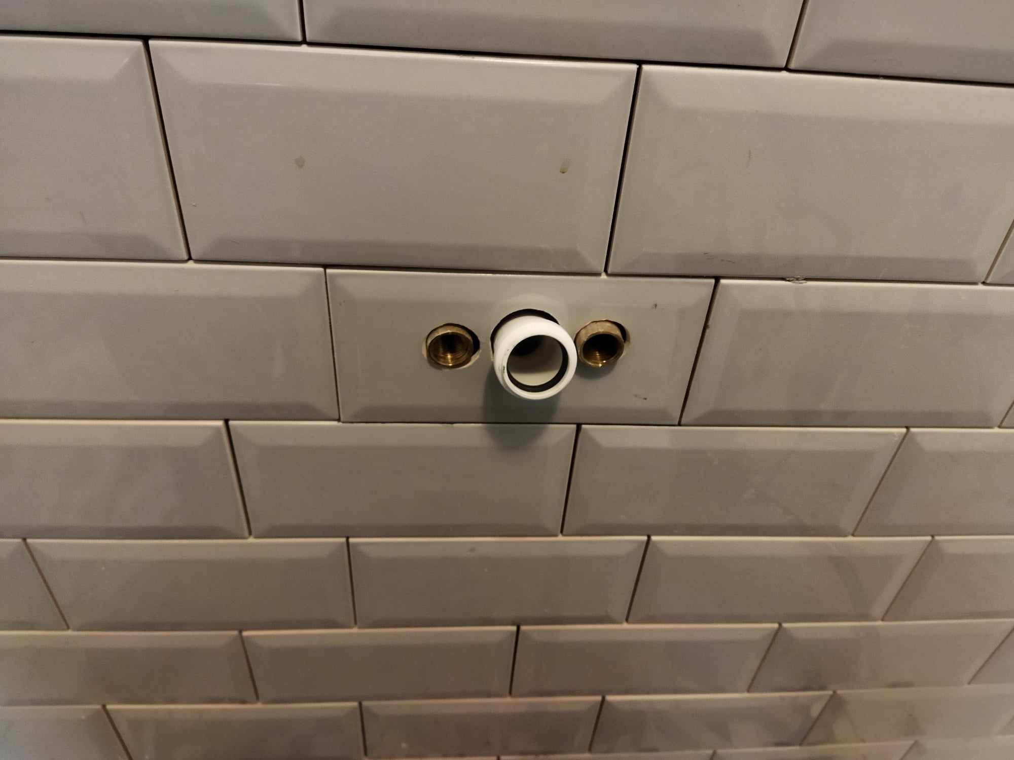 Ściana wyłożona szarymi, prostokątnymi płytkami typu metro z zamontowanymi, mosiężnymi króćcami i białym, plastikowym elementem instalacji wodnej.