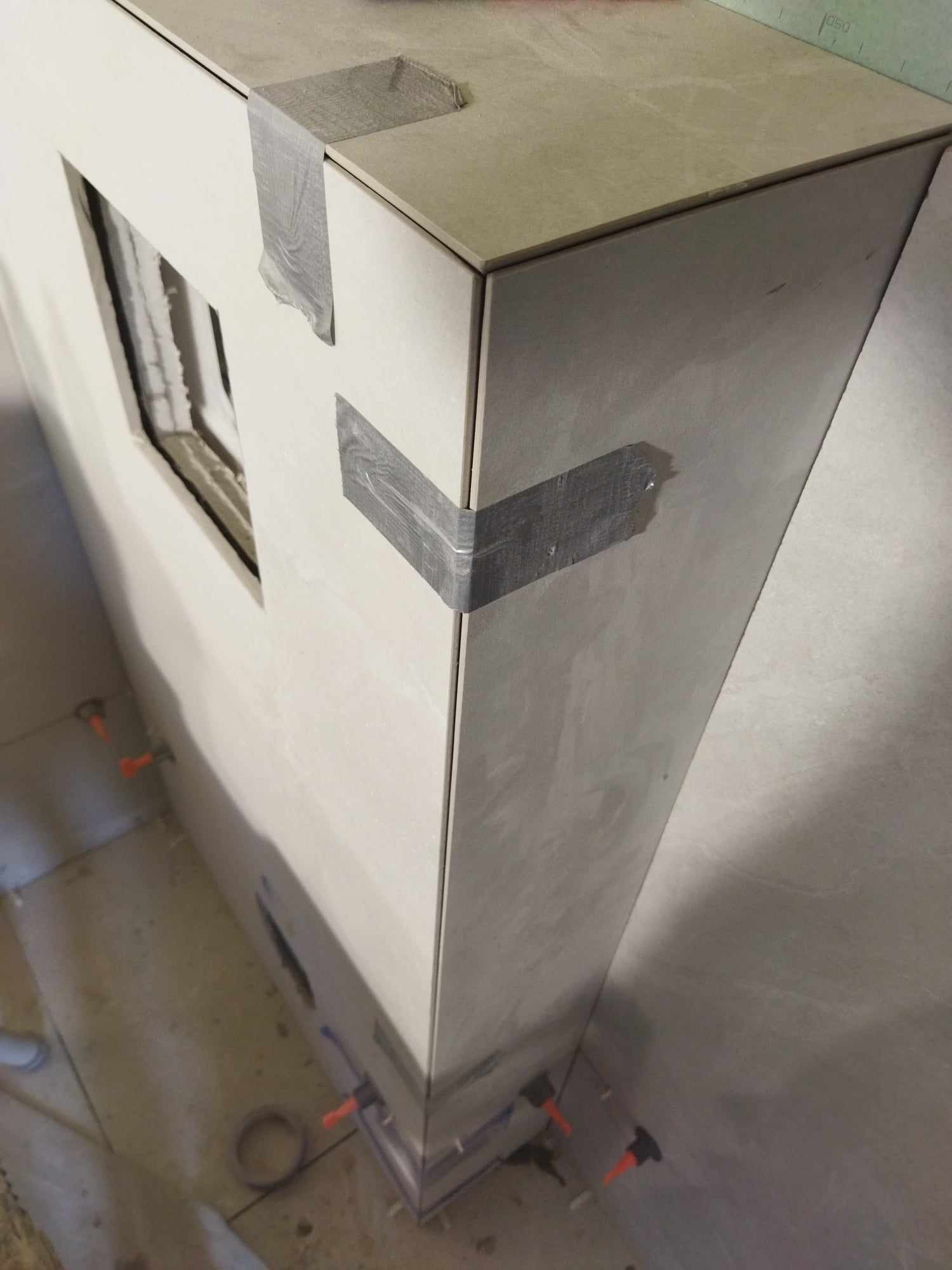 Fragment obudowy z jasnych płytek, zabezpieczonych taśmą malarską na krawędziach, w trakcie prac wykończeniowych, z widocznymi krzyżykami dystansowymi na podłodze.