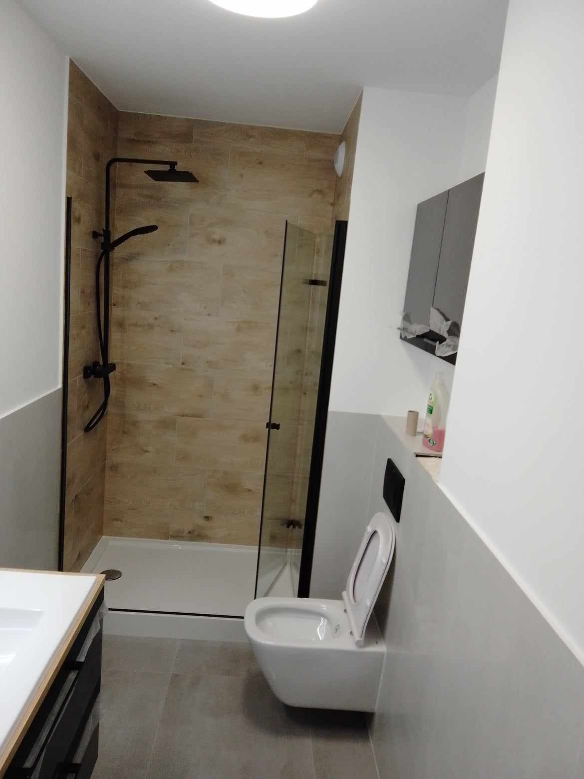 Nowoczesna łazienka po remoncie z kabiną prysznicową typu walk-in z czarnymi ramami i deszczownicą, drewnopodobnymi płytkami na ścianach w strefie prysznica oraz podwieszaną toaletą.