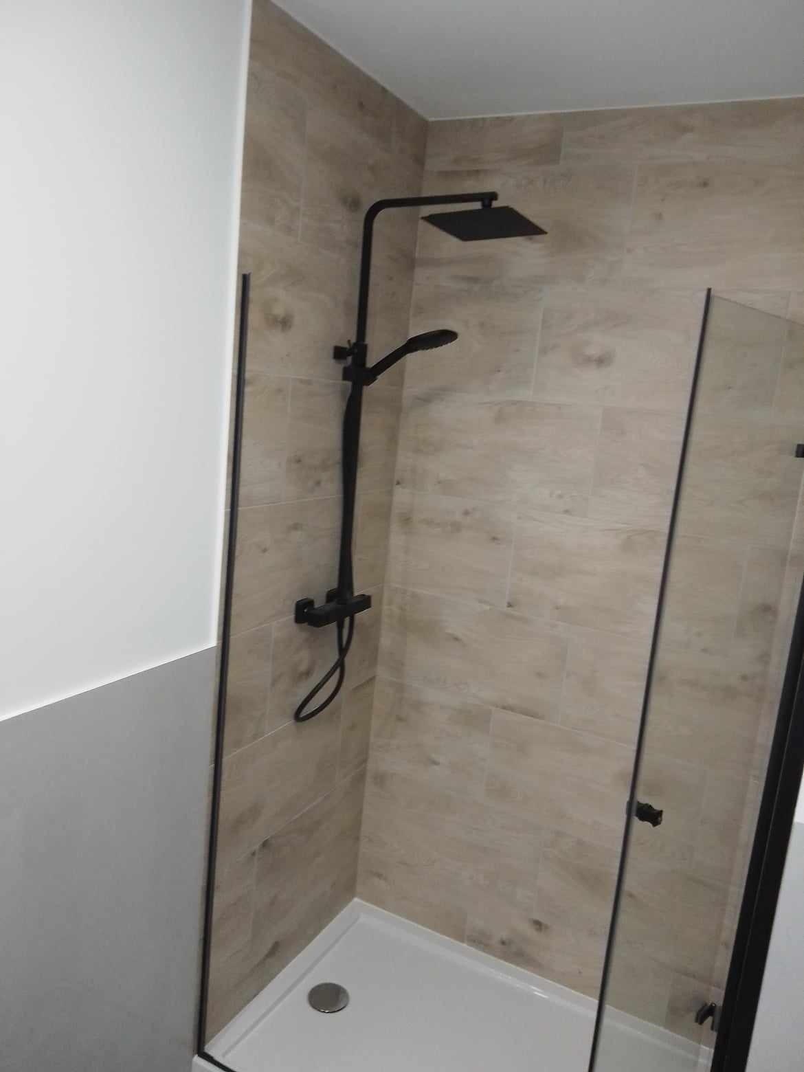 Nowoczesna kabina prysznicowa z czarną armaturą i płytkami imitującymi drewno, widoczny odpływ liniowy i szklane drzwi.