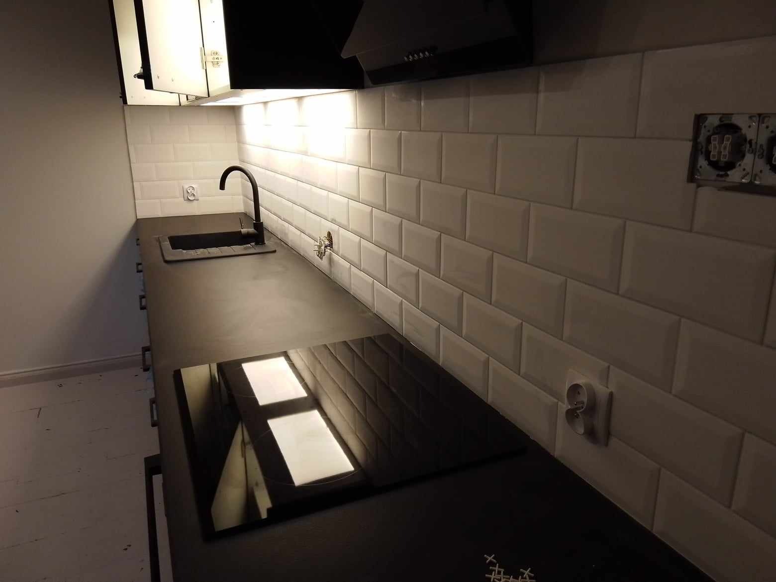 Nowoczesna kuchnia z czarnym blatem, indukcyjną płytą grzewczą i białymi płytkami ułożonymi w cegiełkę. Widoczne gniazdka elektryczne na ścianie i czarna bateria przy zlewie.