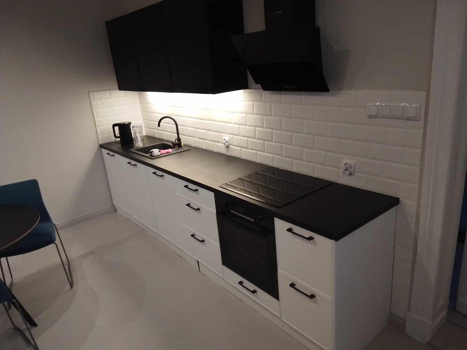 Nowoczesna kuchnia z białymi szafkami, czarnym blatem i płytkami typu metro na ścianie, widoczny czarny okap i płyta indukcyjna.