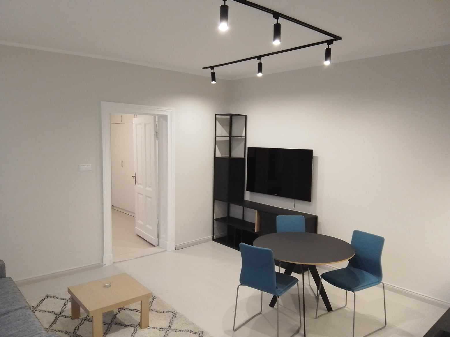 Jasne, pomalowane na szaro mieszkanie z czarnym oświetleniem punktowym, meblami i telewizorem na ścianie. W tle widoczne otwarte drzwi do innego pomieszczenia.