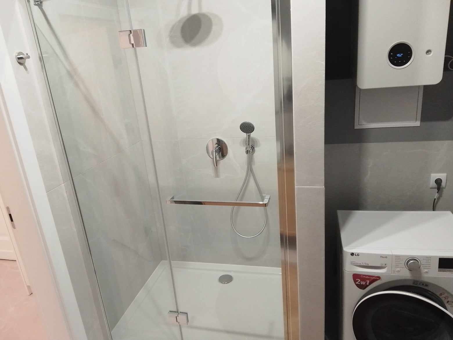 Nowoczesna kabina prysznicowa z szklanymi drzwiami i chromowanymi elementami, obok pralka LG i biały bojler elektryczny na szarej ścianie.