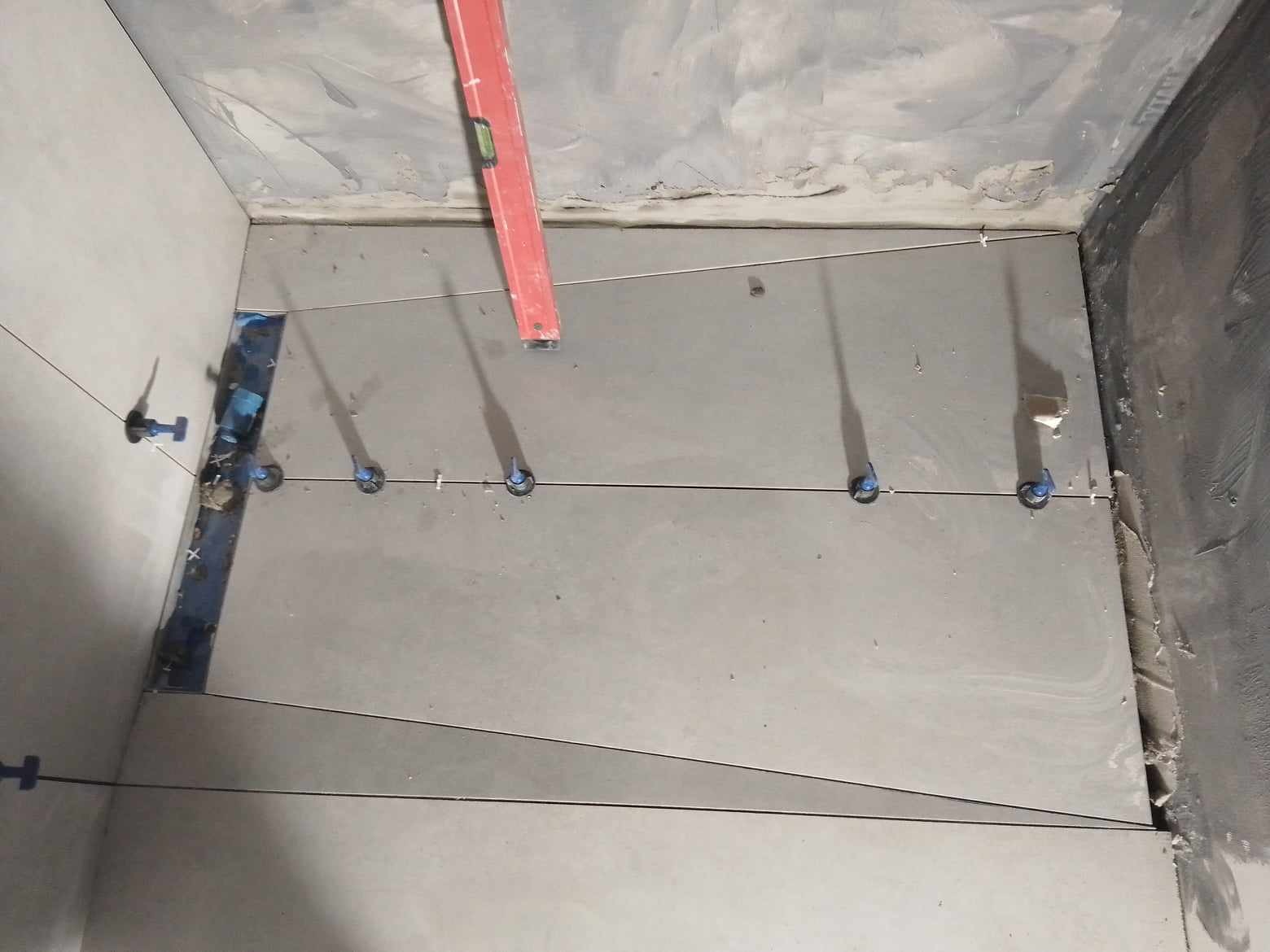 Układanie szarych płytek podłogowych w łazience z użyciem poziomicy i systemu poziomowania płytek, widoczne sznurki wyznaczające linie.