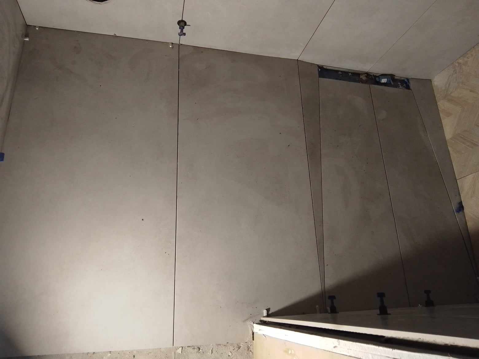 Świeżo ułożone, duże, beżowe płytki na ścianie w łazience, z widocznymi elementami poziomowania i szczelinami dylatacyjnymi, częściowo zakryte folią ochronną, w tle widoczny fragment podłogi...