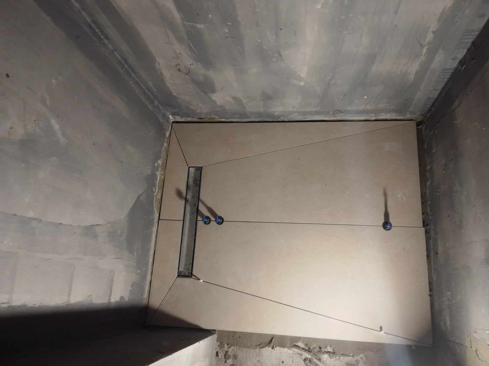 Niedokończona podłoga w łazience z beżowymi płytkami, widoczny odpływ liniowy i elementy poziomujące płytki, surowe ściany w tle.