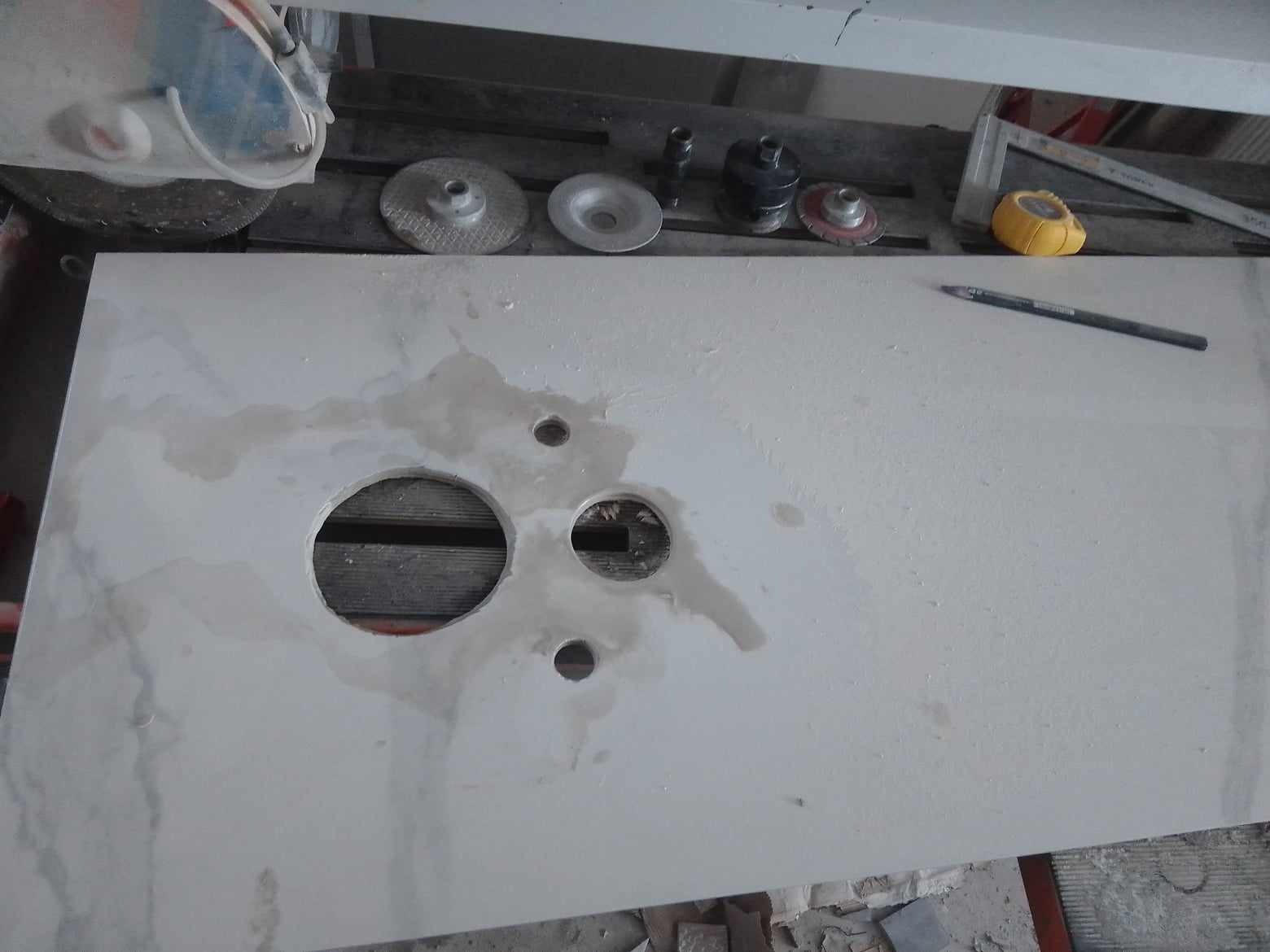 Biały blat z wyciętymi otworami pod umywalkę i armaturę, mokry od wody, narzędzia glazurnicze widoczne w tle.