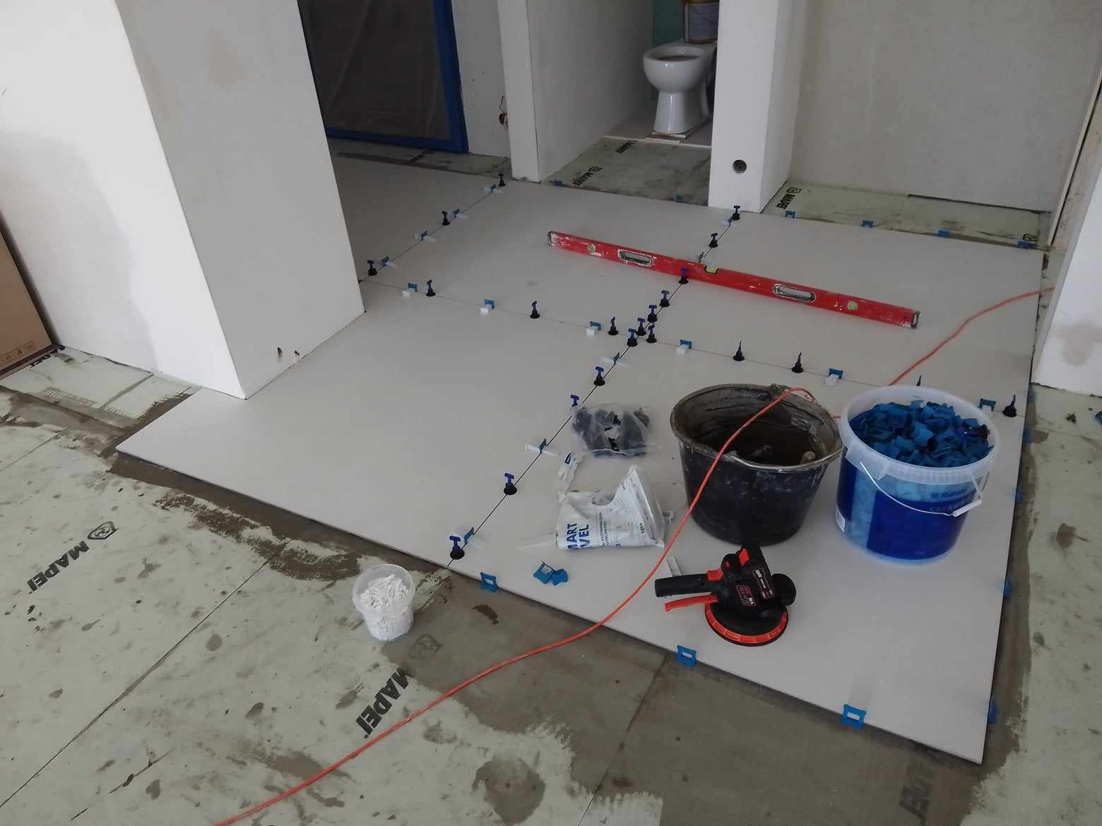 Przygotowanie do ułożenia płytek podłogowych w łazience, widoczne poziomica, przyssawka do płytek, wiadro z zaprawą, wiadro z klipsami poziomującymi i krzyżyki dystansowe.