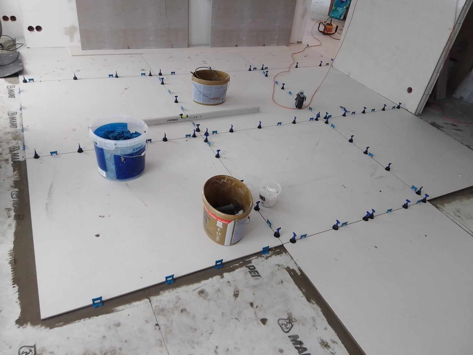 Układanie białych płytek podłogowych z użyciem systemu poziomowania płytek; widoczne wiadra z zaprawą i poziomica.