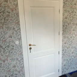 Białe drzwi wewnętrzne z prostokątnymi panelami, złota klamka z kluczem, na tle ściany pokrytej tapetą w motywy leśne ze zwierzętami, widoczny fragment podłogi zabezpieczony folią.