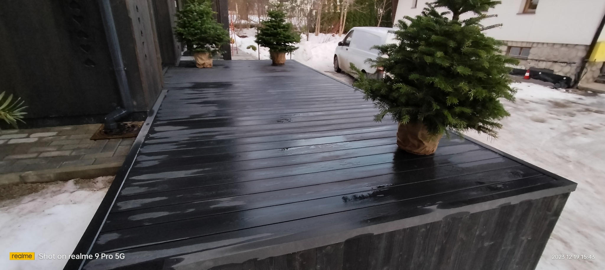 Czarny, drewniany taras z trzema małymi choinkami w jutowych workach, biały samochód dostawczy w tle, śnieg na ziemi.