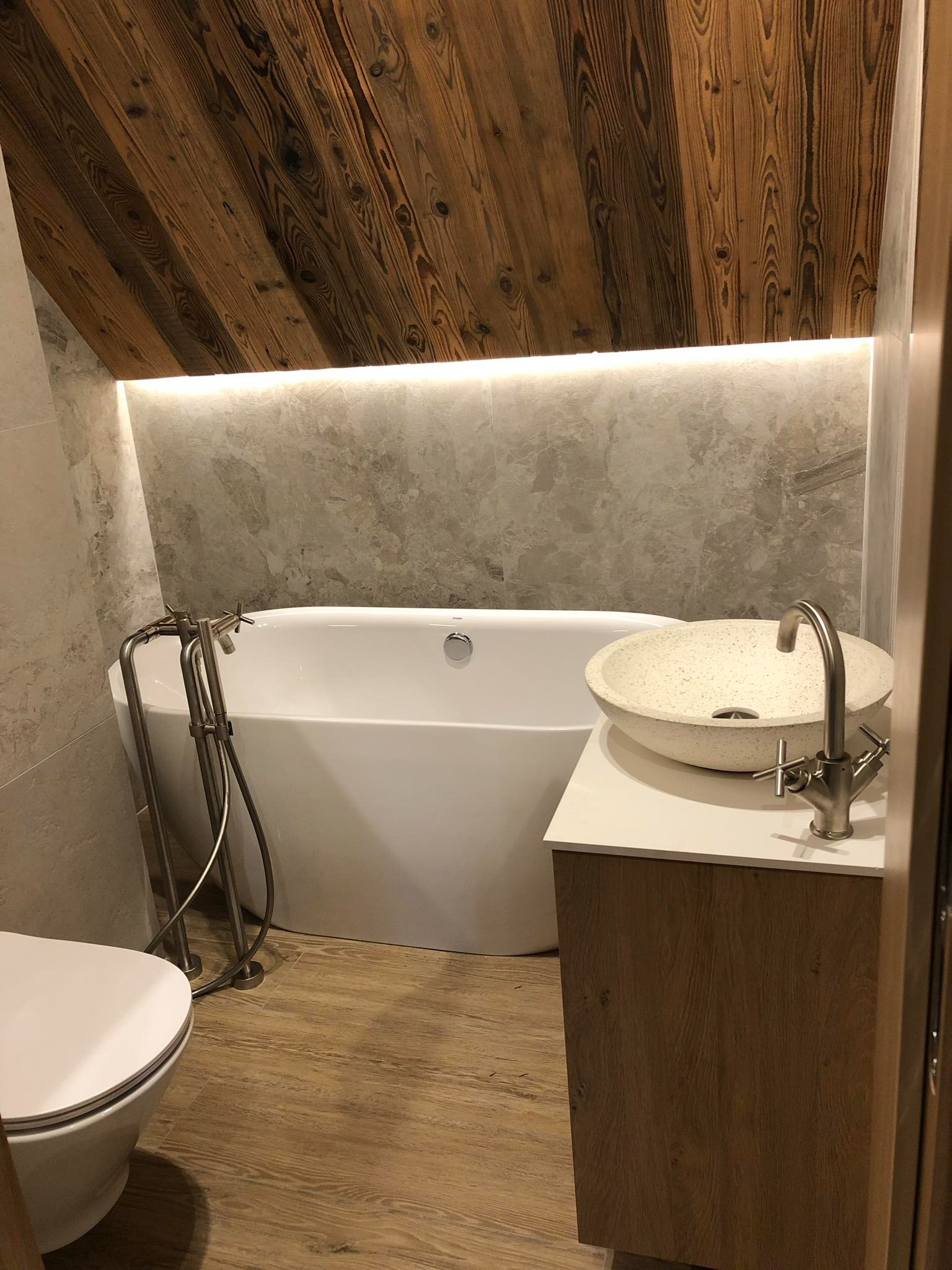 Nowoczesna łazienka na poddaszu z drewnianym sufitem, wolnostojącą wanną, umywalką nablatową w kolorze piaskowym i armaturą w odcieniach srebra.