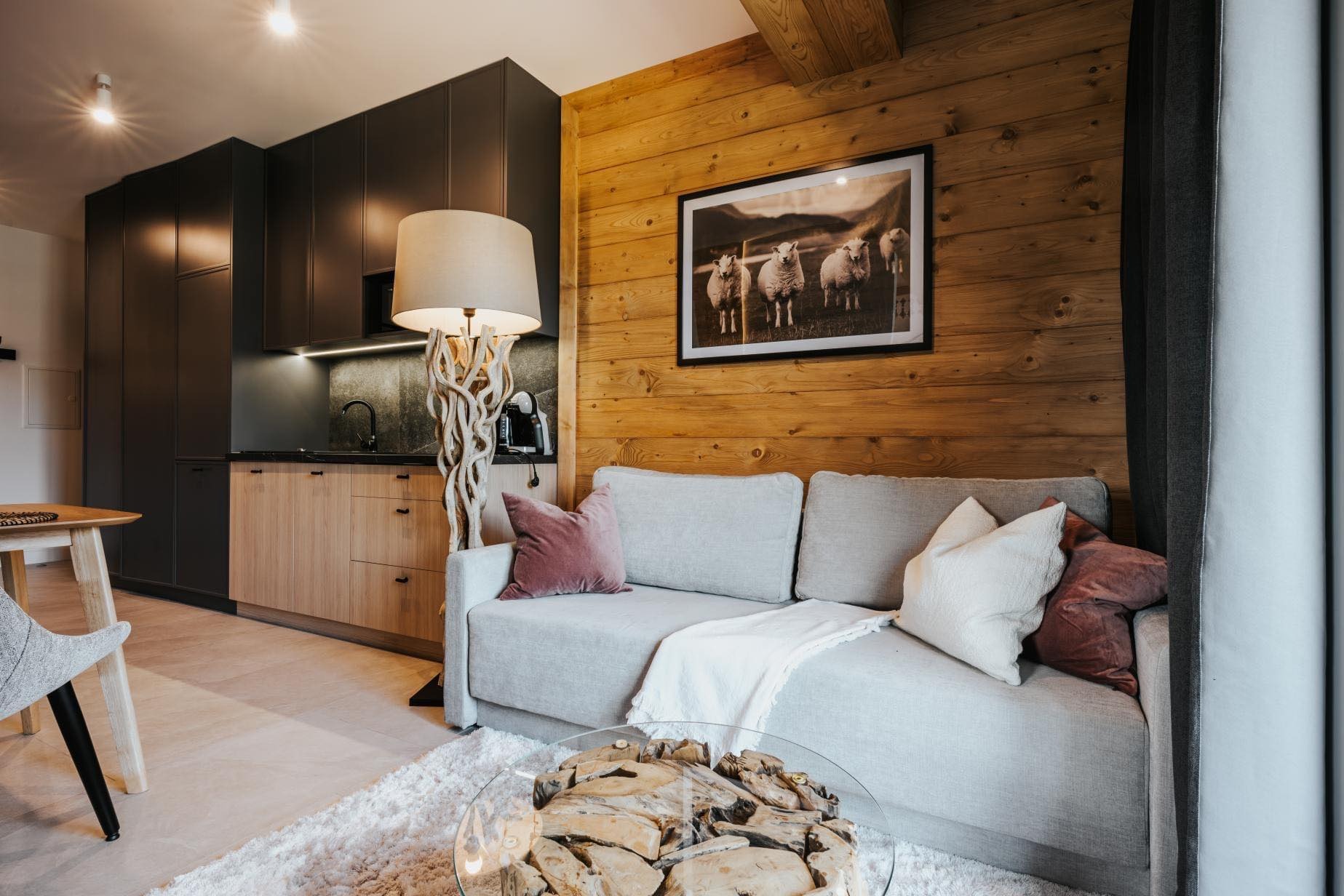 Jasny salon z drewnianą ścianą, szarą sofą z poduszkami, stolikiem kawowym z drewnianymi elementami, lampą z podstawą z gałęzi i aneksem kuchennym w tle z ciemnymi i jasnymi szafkami.