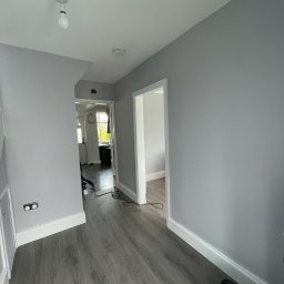 Tomasz Boiński - Wnętrze korytarza po remoncie: świeżo pomalowane szare ściany, ułożone szare panele podłogowe, białe listwy przypodłogowe, widoczne kable i torba narzędziowa.