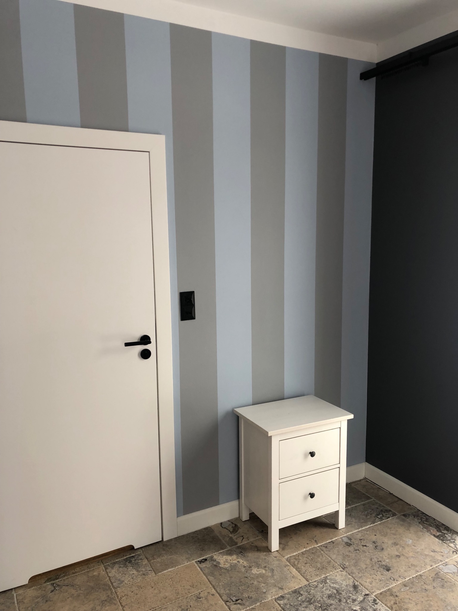 Pokój po odświeżeniu: Ściany w pionowe pasy w odcieniach szarości i błękitu, biała szafka nocna z czarnymi gałkami, białe drzwi z czarną klamką i ciemnoszara ściana w rogu, na podłodze płytki...