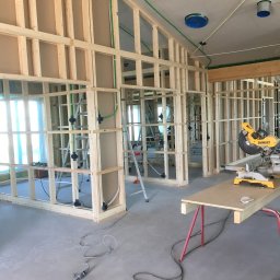 Kamil Muranski - Wnętrze domu w trakcie budowy, widoczne drewniane ramy ścian działowych, instalacje elektryczne w zielonych rurach, drabina, piła ukośnica na stole roboczym, surowy beton na podłodze...