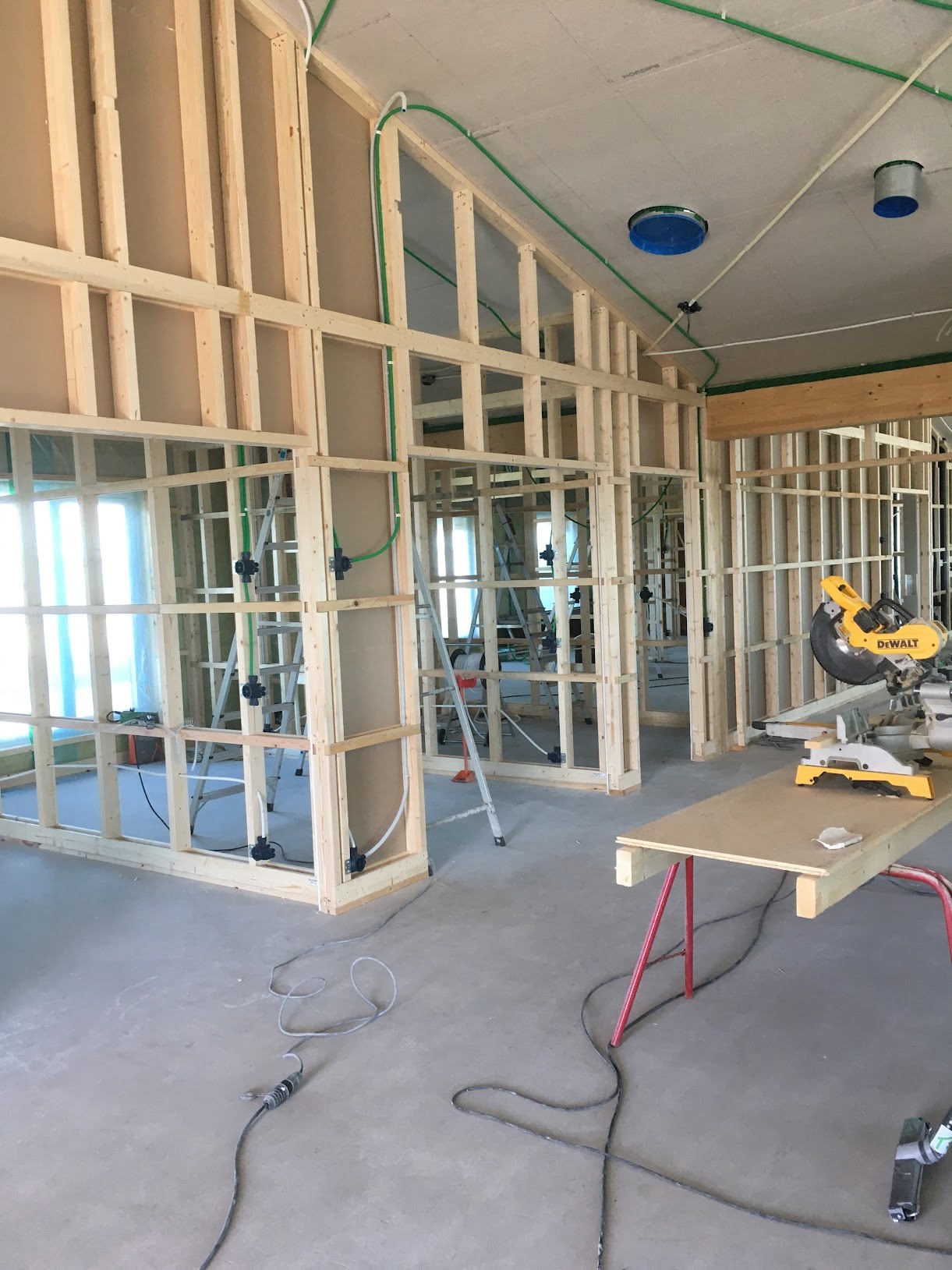Wnętrze domu w trakcie budowy, widoczne drewniane ramy ścian działowych, instalacje elektryczne w zielonych rurach, drabina, piła ukośnica na stole roboczym, surowy beton na podłodze...