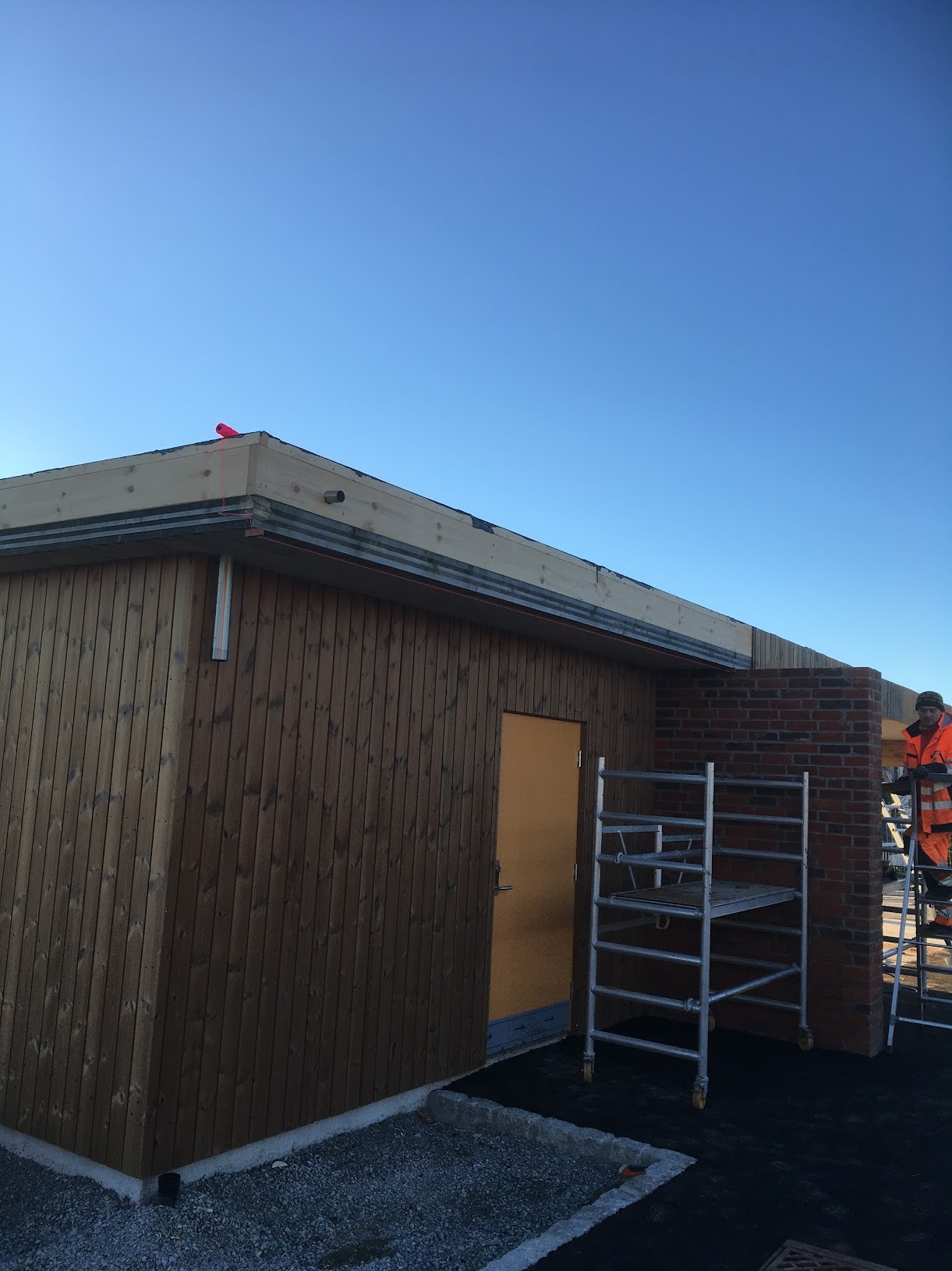 Budynek w trakcie budowy z drewnianą elewacją, widoczne rusztowanie, ceglana ściana i pracownik w pomarańczowym kombinezonie na tle błękitnego nieba.