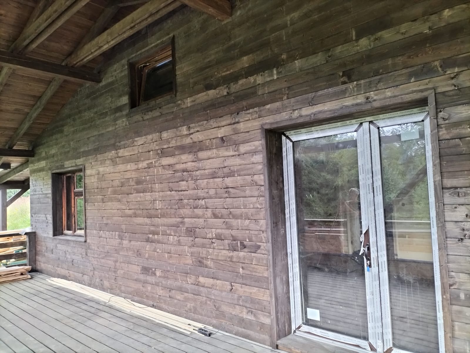 Drewniana elewacja domu z widocznymi oknami i drzwiami balkonowymi z białymi ramami, na podłodze leżą listwy przypodłogowe.