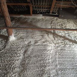 DOM BEZ WAD SPÓŁKA Z OGRANICZONĄ ODPOWIEDZIALNOŚCIĄ - Poddasze po ociepleniu pianką: widoczna struktura piany na ścianach i drewnianych elementach konstrukcyjnych, z bagażem i kartonem w tle.