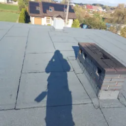 Szary dach pokryty papą termozgrzewalną, widoczny komin z cegły klinkierowej i cień osoby robiącej zdjęcie, w tle dom z panelami słonecznymi.