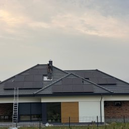 DOM BEZ WAD SPÓŁKA Z OGRANICZONĄ ODPOWIEDZIALNOŚCIĄ - Montaż paneli słonecznych na dachu domu jednorodzinnego z wykorzystaniem drabiny, widoczny pracownik w trakcie instalacji.