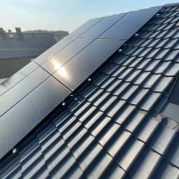 DOM BEZ WAD SPÓŁKA Z OGRANICZONĄ ODPOWIEDZIALNOŚCIĄ - Panele słoneczne zamontowane na ciemnoszarym dachu z dachówki ceramicznej, widoczne elementy montażowe, w tle fragment dachu sąsiedniego budynku i zarys drzew.