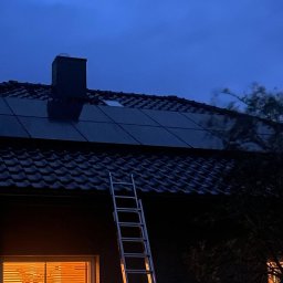 DOM BEZ WAD SPÓŁKA Z OGRANICZONĄ ODPOWIEDZIALNOŚCIĄ - Panel słoneczny na dachu domu z drabina opartą o ścianę, widok wieczorową porą, ciepłe światło z okna.