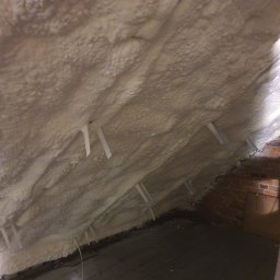 DOM BEZ WAD SPÓŁKA Z OGRANICZONĄ ODPOWIEDZIALNOŚCIĄ - Poddasze ocieplone pianką PUR, widoczne drewniane deski podłogowe i fragment ściany z cegły, z plastikowymi elementami mocującymi piankę.