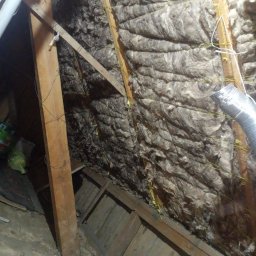 DOM BEZ WAD SPÓŁKA Z OGRANICZONĄ ODPOWIEDZIALNOŚCIĄ - Poddasze z drewnianą konstrukcją i izolacją z wełny mineralnej, zabezpieczoną sznurkiem. Widoczny fragment rury wentylacyjnej i resztki materiałów budowlanych na podłodze.