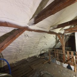 DOM BEZ WAD SPÓŁKA Z OGRANICZONĄ ODPOWIEDZIALNOŚCIĄ - Poddasze z drewnianą konstrukcją pokryte grubą warstwą jasnej pianki izolacyjnej, widoczne metalowe elementy mocujące, drewniany pomost i worki z odpadami na podłodze.