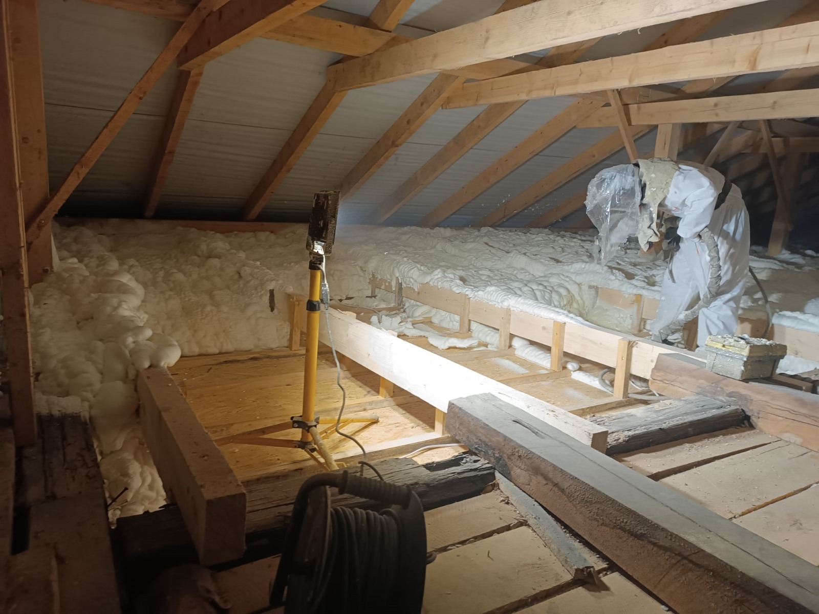 Pracownik w kombinezonie ochronnym aplikuje piankę izolacyjną na poddaszu, widoczna drewniana konstrukcja dachu i oświetlenie robocze.
