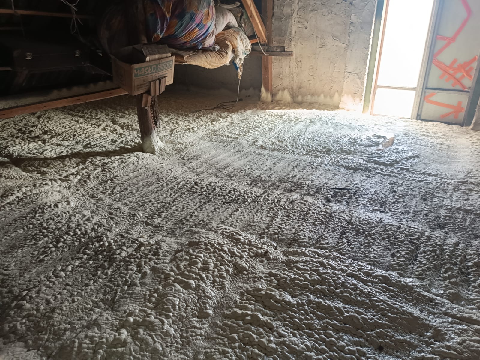 Poddasze po natrysku izolacji piankowej PUR, widoczne ślady po rozpylaniu, kartonowe pudełko przymocowane do drewnianego słupa, jasne światło wpadające przez otwór drzwiowy.