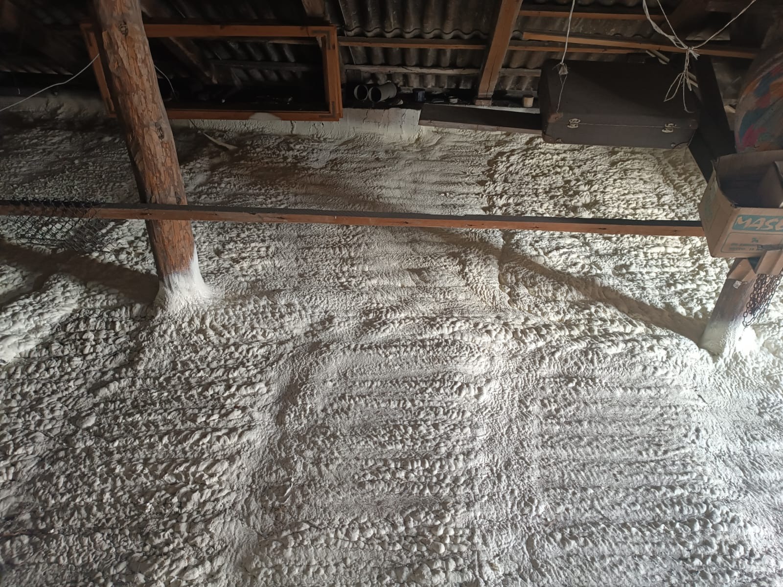 Poddasze po ociepleniu pianką: widoczna struktura piany na ścianach i drewnianych elementach konstrukcyjnych, z bagażem i kartonem w tle.