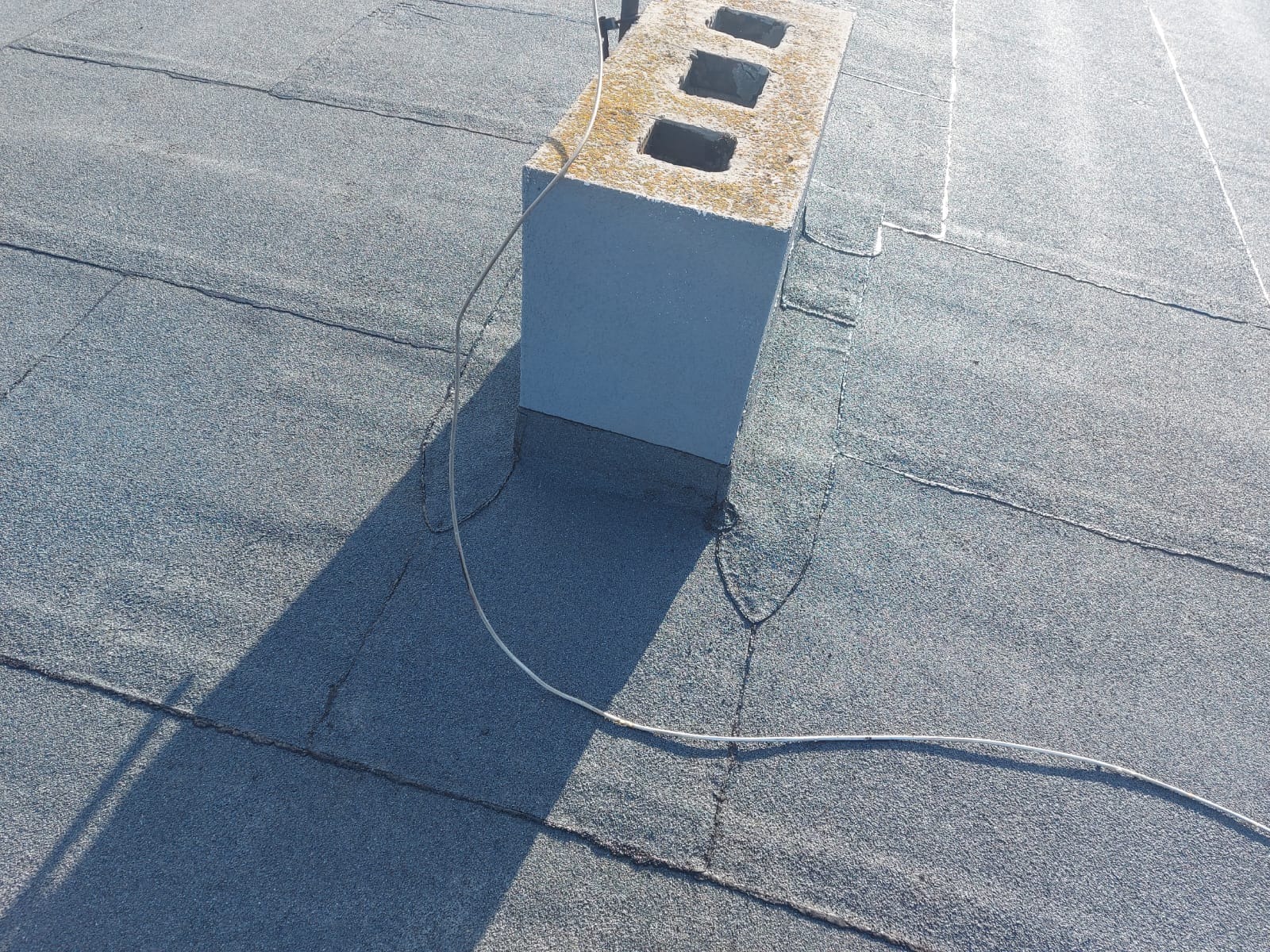 Szary, płaski dach pokryty papą z widocznym kominem wentylacyjnym z trzema otworami i przewodem elektrycznym. Widoczne łączenia papy i cień komina.