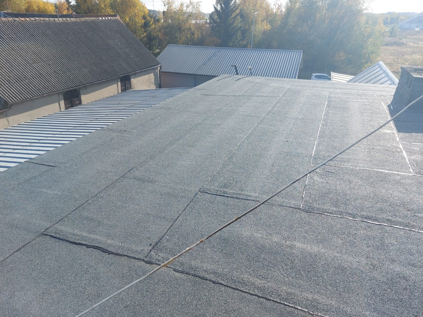 Widok z góry na płaski dach pokryty papą termozgrzewalną, z widocznymi łączeniami i elementami konstrukcyjnymi, w tle budynki gospodarcze o różnych pokryciach dachowych i jesienne drzewa.