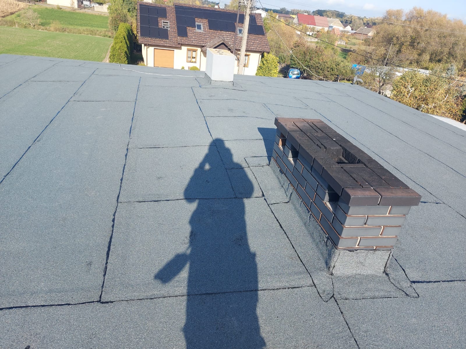 Szary dach pokryty papą termozgrzewalną, widoczny komin z cegły klinkierowej i cień osoby robiącej zdjęcie, w tle dom z panelami słonecznymi.