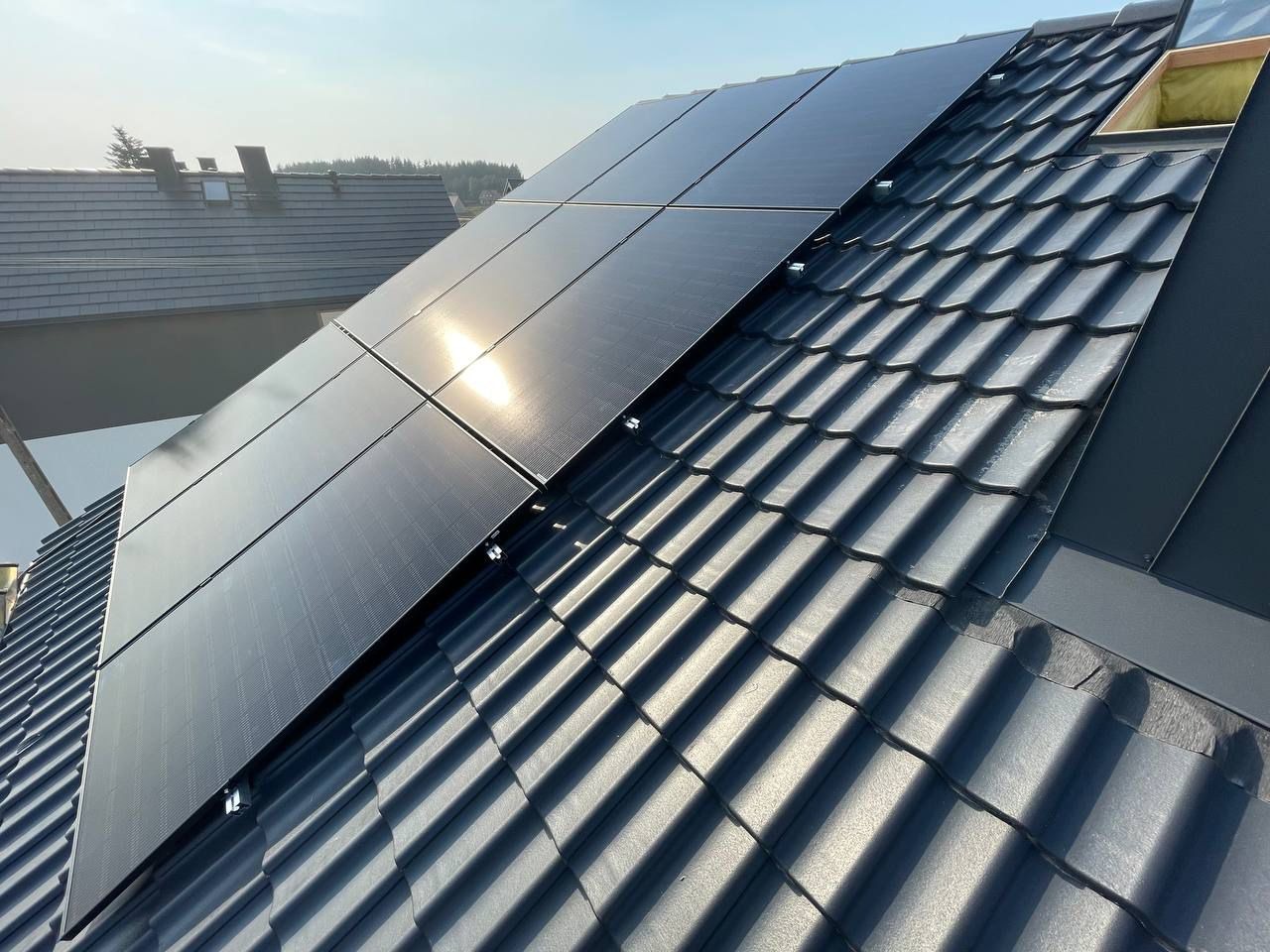 Panele słoneczne zamontowane na ciemnoszarym dachu z dachówki ceramicznej, widoczne elementy montażowe, w tle fragment dachu sąsiedniego budynku i zarys drzew.