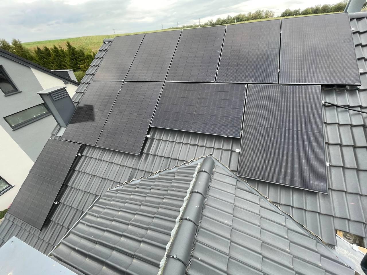 Panele słoneczne zamontowane na szarym dachu domu jednorodzinnego, widok z góry, w tle pole i las